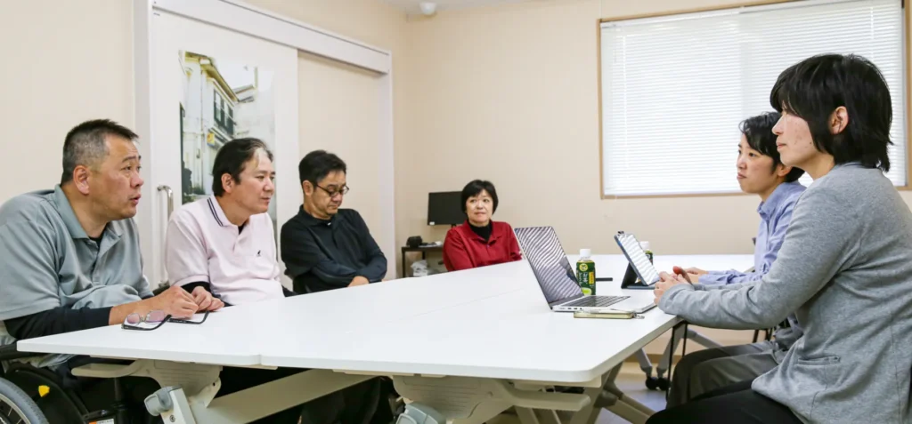 pic--01 - ホームページ作成ならL-planningへ Six colleagues sit around a white conference table in a meeting room, discussing with laptops and tablets on the table.