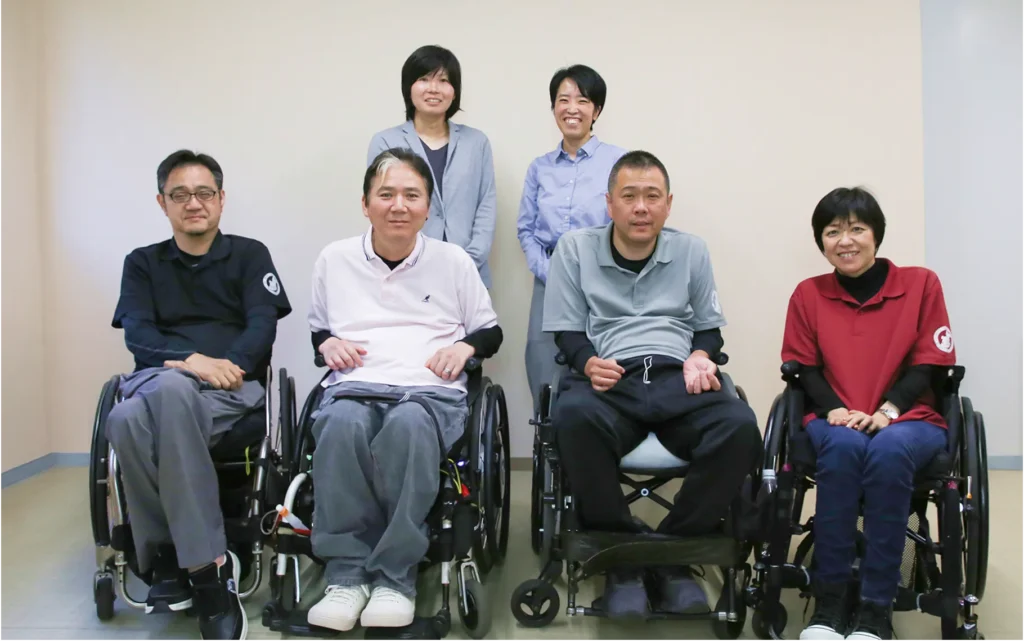 pic--main - ホームページ作成ならL-planningへ Group photo of six adults in a room: four in wheelchairs in the front row and two standing behind, all smiling at the camera.