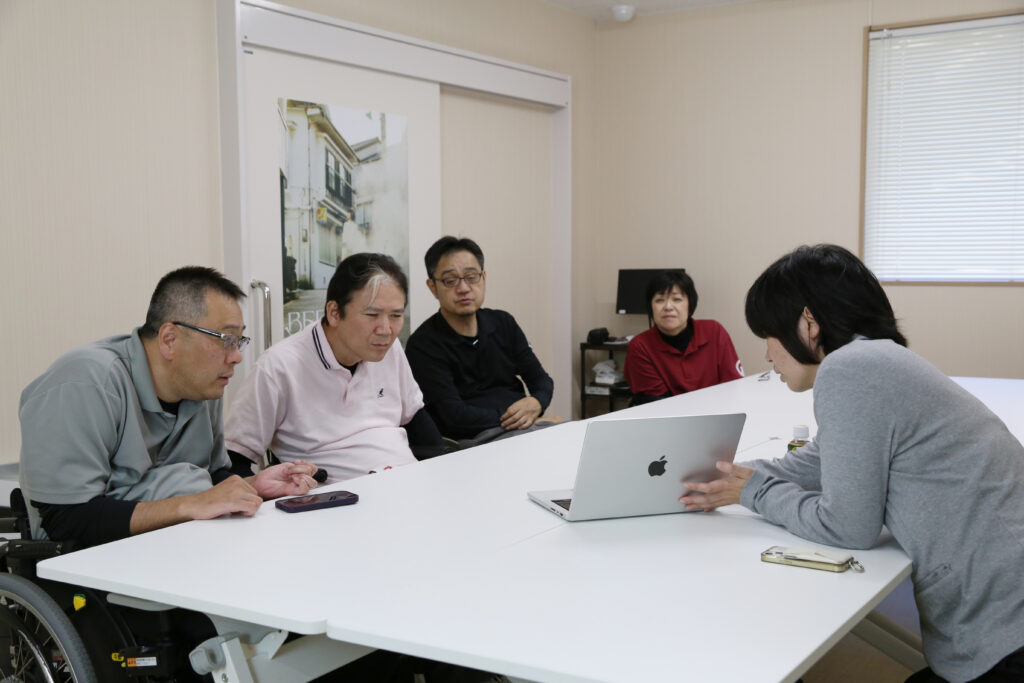 IMG_3521調整 - ホームページ作成ならL-planningへ Group of five adults around a white conference table; one person in a wheelchair, laptops in use, in a bright office.